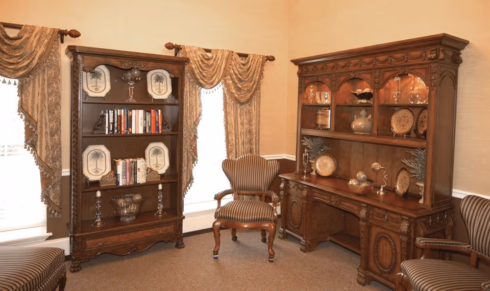 Warmly lit sitting room with carved wooden bookcases and cabinets, decorative plates and vases, and striped upholstered chairs.