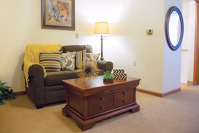 A cozy living room area with a dark green loveseat adorned with multiple patterned pillows and a yellow throw blanket. In front of the loveseat is a wooden coffee table with decorative items including a spiky sculpture, a small green plant, and a black and white chevron-patterned box. A floor lamp with a beige shade stands behind the loveseat, and a framed floral artwork hangs on the wall above. The room has beige carpeting and light-colored walls, with a round mirror visible on a nearby wall.