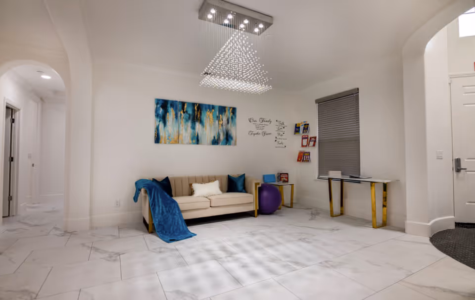 A bright and modern living room area with white walls and large white tiled floor. There is a beige sofa with blue and white pillows and a blue throw blanket. Above the sofa is a blue and gold abstract painting. To the right of the sofa is a purple exercise ball and a small side table with a blue laptop. On the wall behind the sofa are inspirational wall decals and a rack holding colorful brochures. A window with closed blinds is next to a white desk with gold legs. The room is illuminated by a contemporary ceiling light fixture with hanging crystals. There are two archways visible, one leading to a hallway and the other near the entrance door.