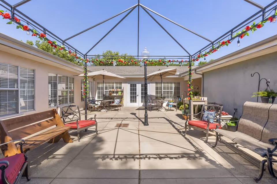 Sunny enclosed courtyard with seating, tables, umbrellas, and a decorative metal pergola strung with flowers.