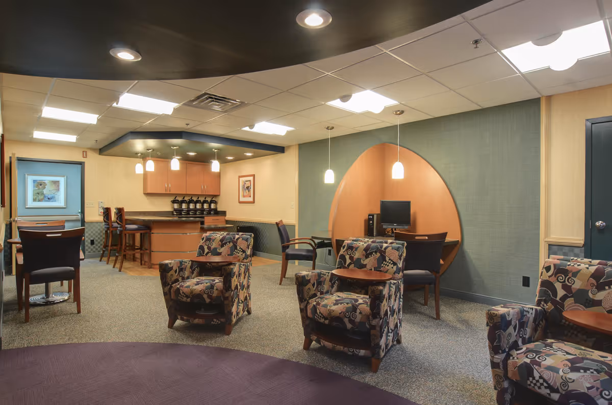 A cozy common area in a senior living facility featuring patterned armchairs with attached small tables, a computer workstation set into a wooden alcove on the right wall, a small bar area with high chairs and coffee dispensers in the background, and soft overhead lighting.