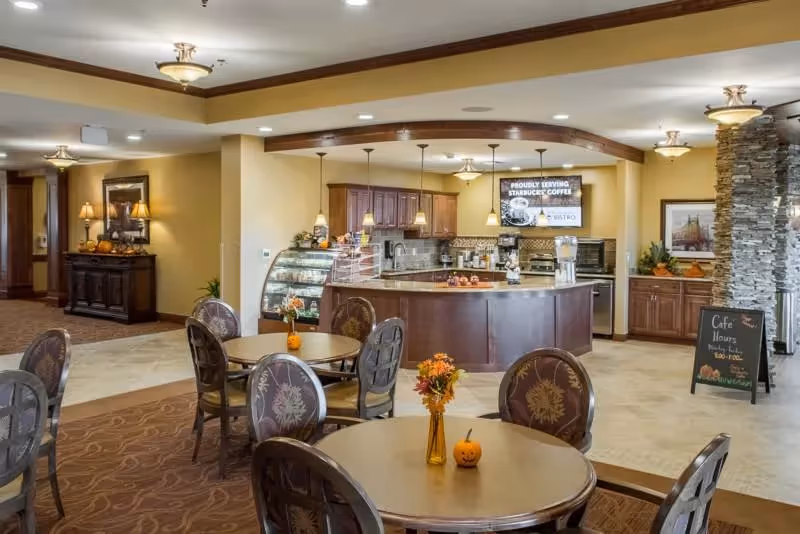 Cafe-style dining area with round tables and chairs facing a service counter, display case, and coffee station inside a senior living facility.