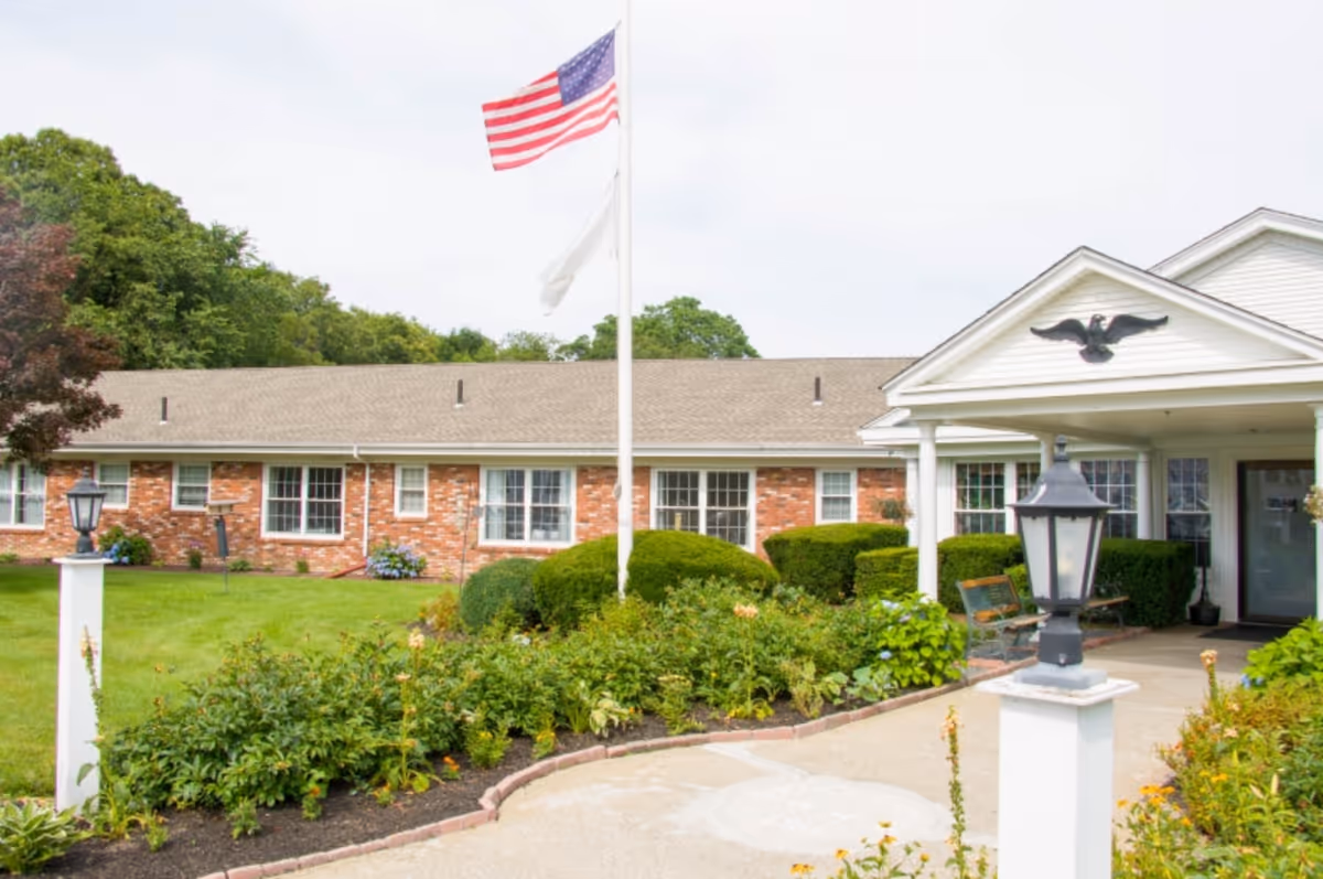 Exterior view of a single-story brick building with white trim, a covered entrance with columns, and an American flag on a flagpole in front. The area is landscaped with green bushes, plants, and a well-maintained lawn.