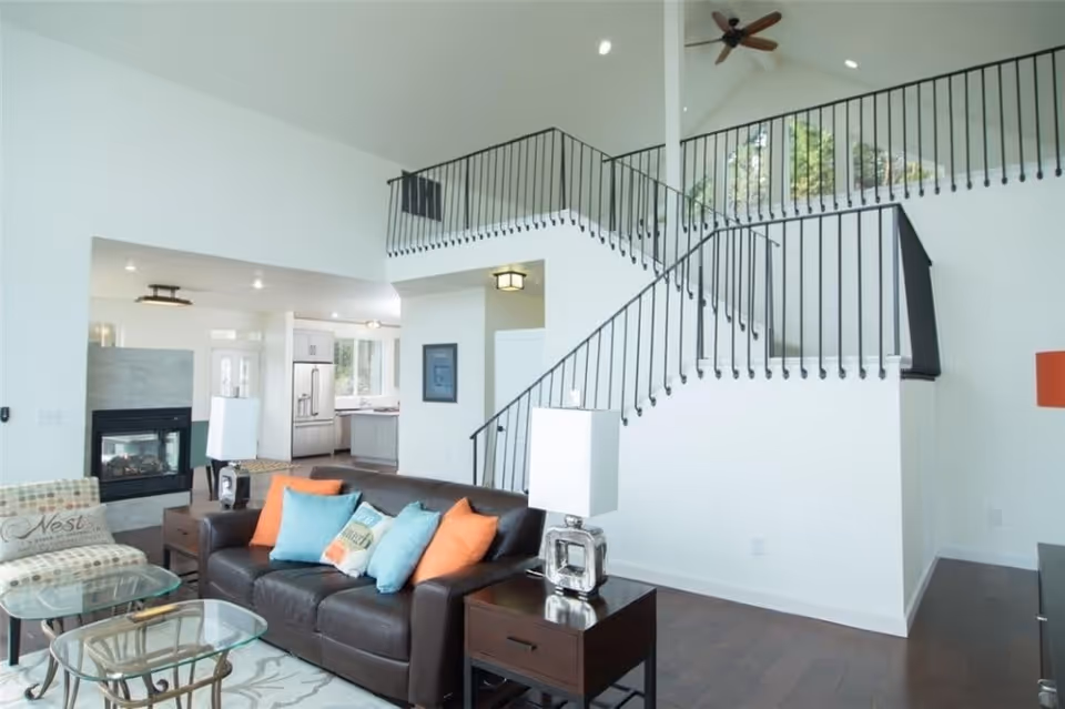 Bright and spacious living room with a dark brown leather sofa adorned with blue and orange pillows, a patterned armchair, glass coffee tables, a fireplace, and a staircase with black metal railings leading to an upper floor. The room has white walls, dark wood flooring, and large windows letting in natural light.