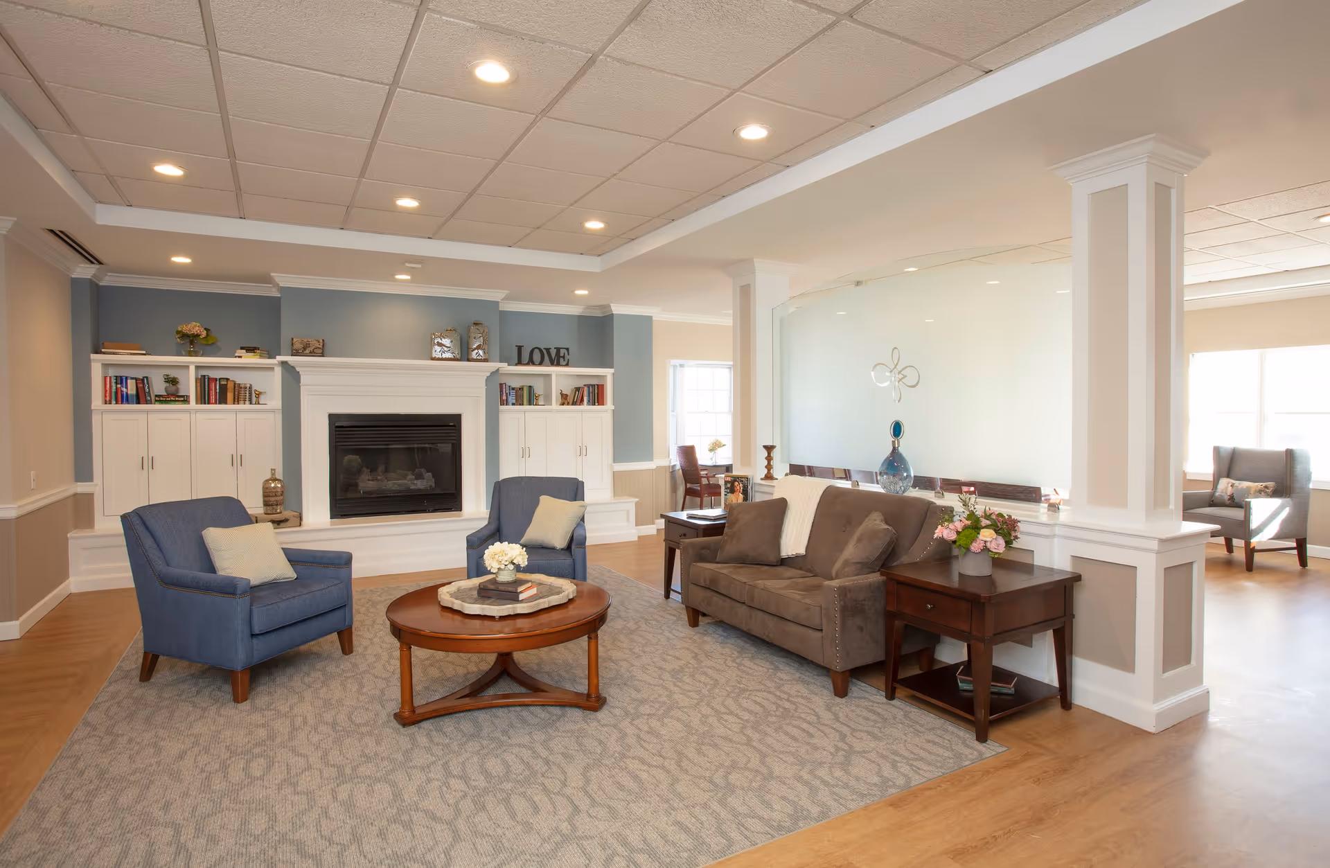 A cozy assisted living facility common area with a brown sofa, two blue armchairs, a round wooden coffee table with books and flowers, built-in white shelves with books and decor, a fireplace, and a decorative sign that says 'LOVE'. The room has soft lighting, a patterned carpet, and wood flooring.