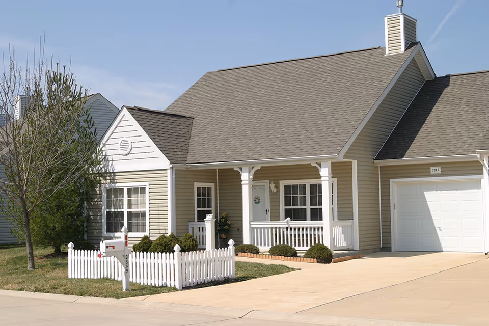 Exterior view of a single-story house with beige siding, a gray shingled roof, a white picket fence around a small front yard, a driveway leading to a white garage door, and a covered front porch with white railings and columns.