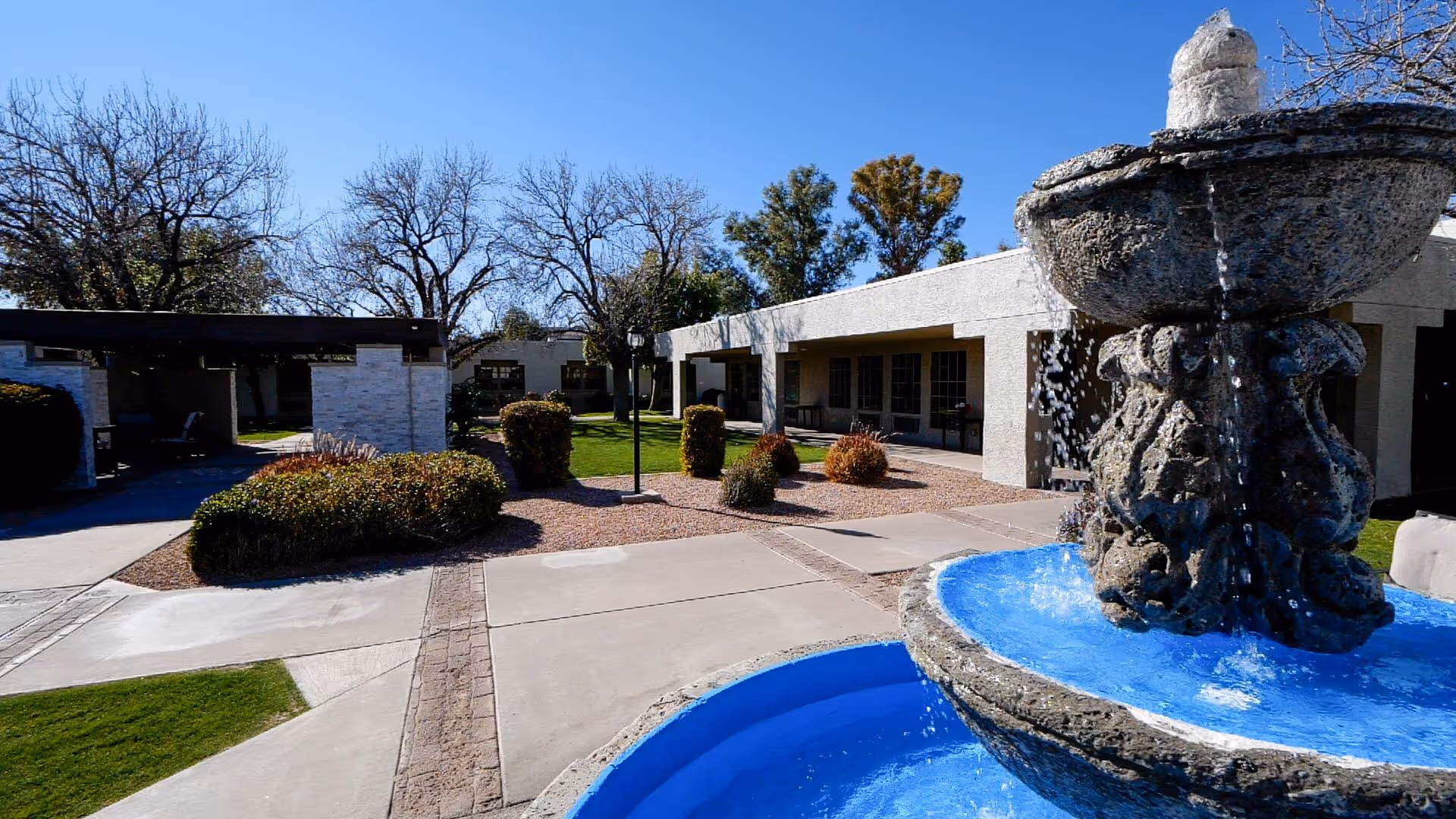Outdoor courtyard area of a senior living facility with a stone water fountain in the foreground, surrounded by paved walkways, green grass, bushes, and trees with leafless branches. Single-story buildings with covered patios are visible in the background under a clear blue sky.