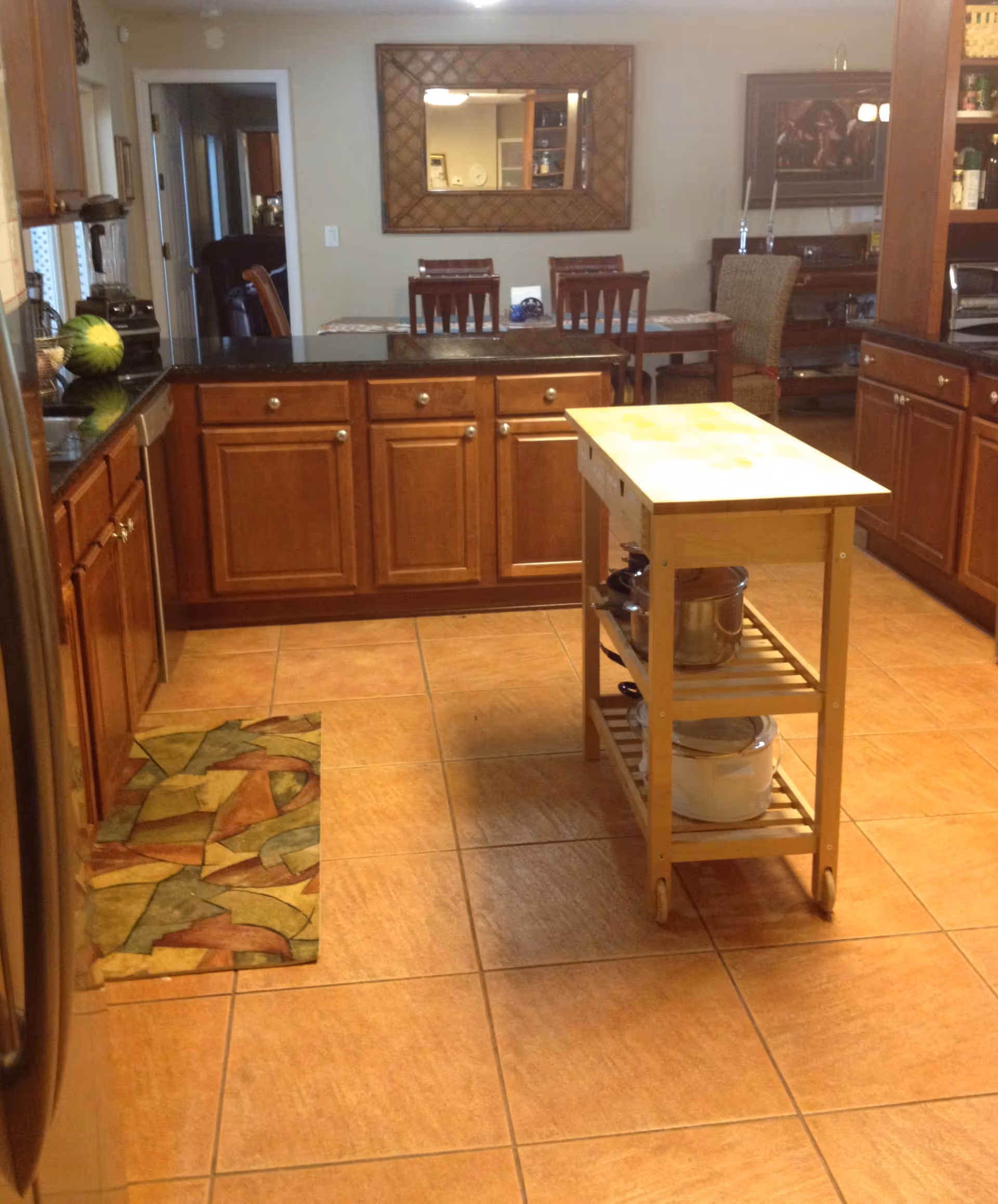 A kitchen area with wooden cabinets and black countertops. There is a small wooden kitchen island with shelves holding pots and pans in the center. A colorful patterned rug is placed on the tiled floor near the sink. In the background, a dining table with four chairs is visible, along with a large decorative mirror on the wall and some additional furniture and decor.