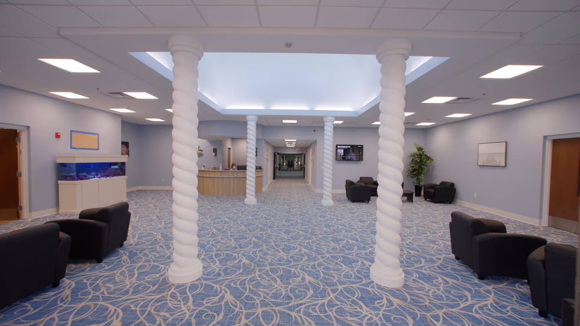 A spacious, well-lit common area with blue patterned carpet and light blue walls. The room features four white twisted columns supporting a recessed ceiling with soft lighting. There are several black armchairs arranged along the sides, a large fish tank on the left, a reception desk in the background, a wall-mounted TV, and a potted plant on the right.