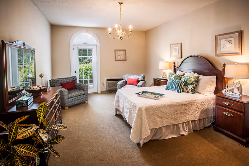 A cozy bedroom with a large bed covered in a white quilt and multiple decorative pillows. There are two wooden nightstands with lamps on either side of the bed. Two patterned armchairs with red cushions are placed near a glass door with an arched window above it, letting in natural light. A wooden dresser with a mirror and decorative items is on the left side of the room. A green leafy plant is visible in the foreground.
