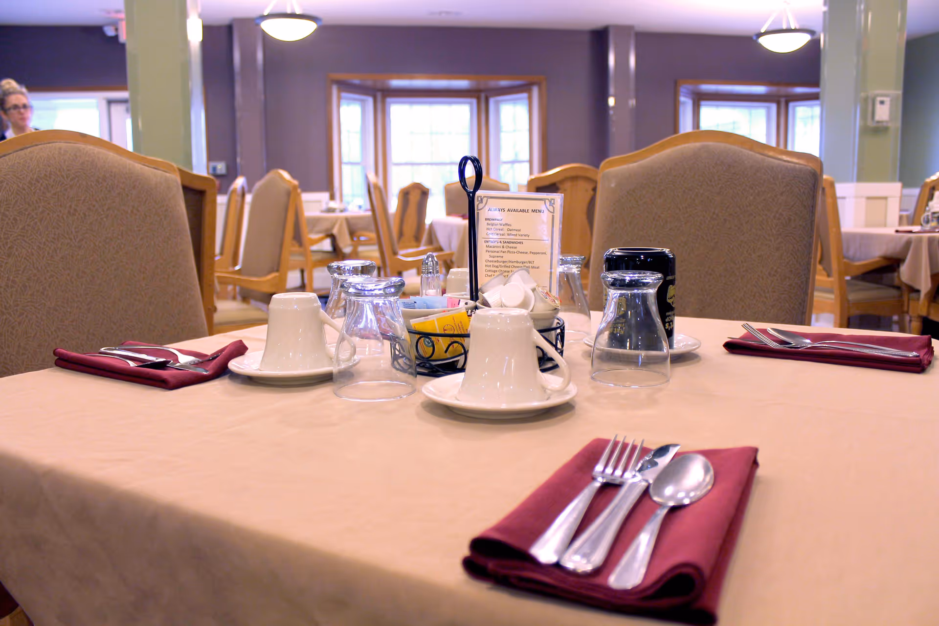 A dining table set for four in a dining room with beige tablecloth and maroon napkins. Each place setting includes a fork, knife, and spoon on a folded napkin, along with an upside-down glass and a white cup on a saucer. The table also has a small basket with sugar packets, creamers, salt and pepper shakers, and a menu card holder. The background shows more tables and chairs, large windows, and soft lighting.