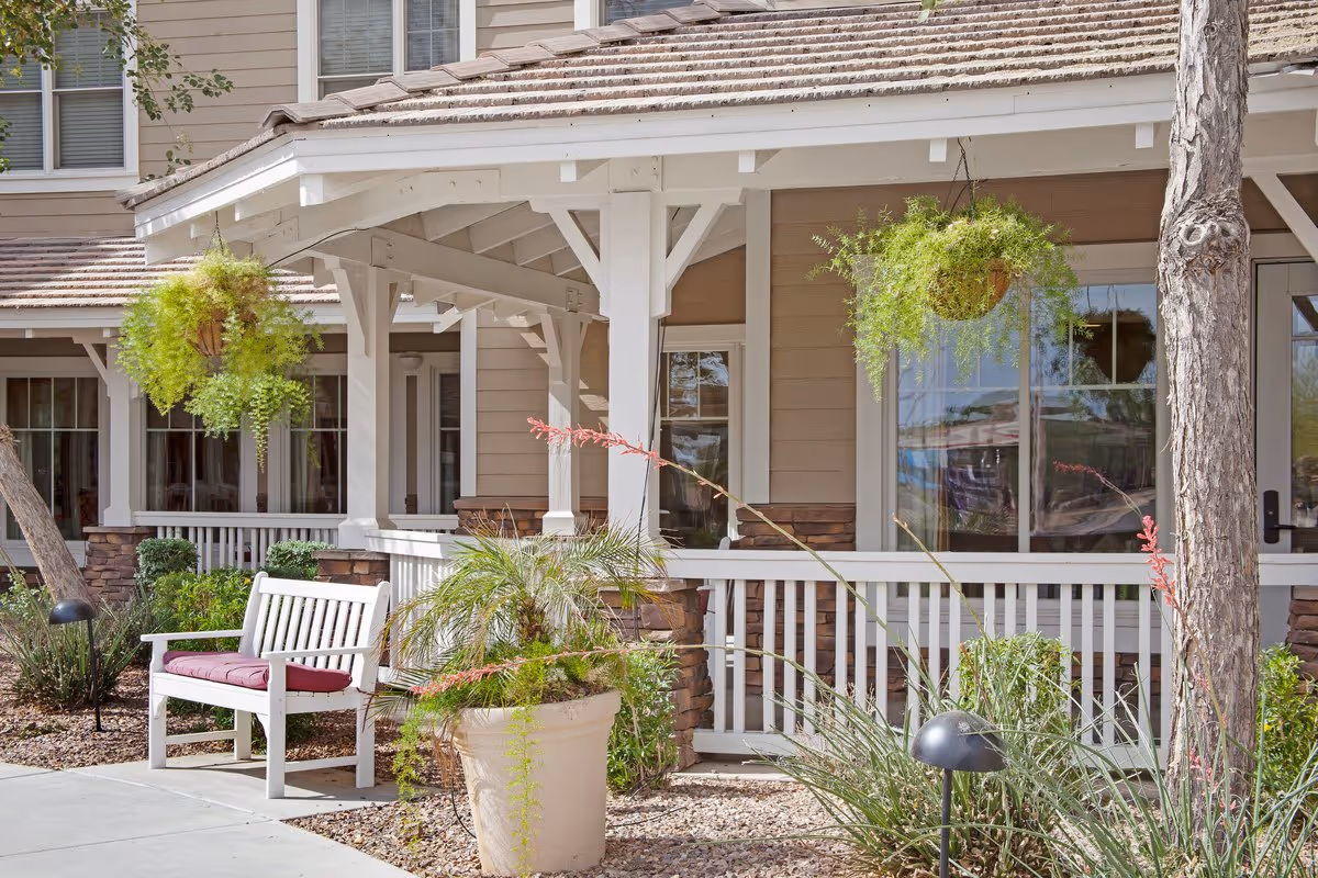 Covered front porch of a senior living residence featuring a white bench with a cushion, hanging potted ferns, a large planter, and a railing.