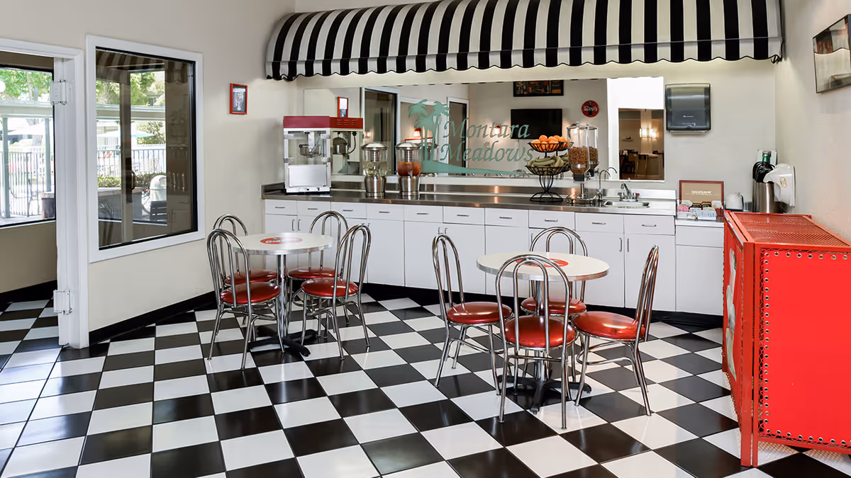 A retro-style dining area with black and white checkered floor tiles, two round tables each surrounded by four metal chairs with red cushioned seats. The back wall features a long counter with white cabinets, a popcorn machine, beverage dispensers, a fruit basket, and a cereal dispenser. Above the counter is a large mirror with the words 'Montara Meadows' and a palm tree graphic. A black and white striped awning is mounted above the mirror. There is a red vending machine on the right side and a window and door on the left side showing an outdoor area.