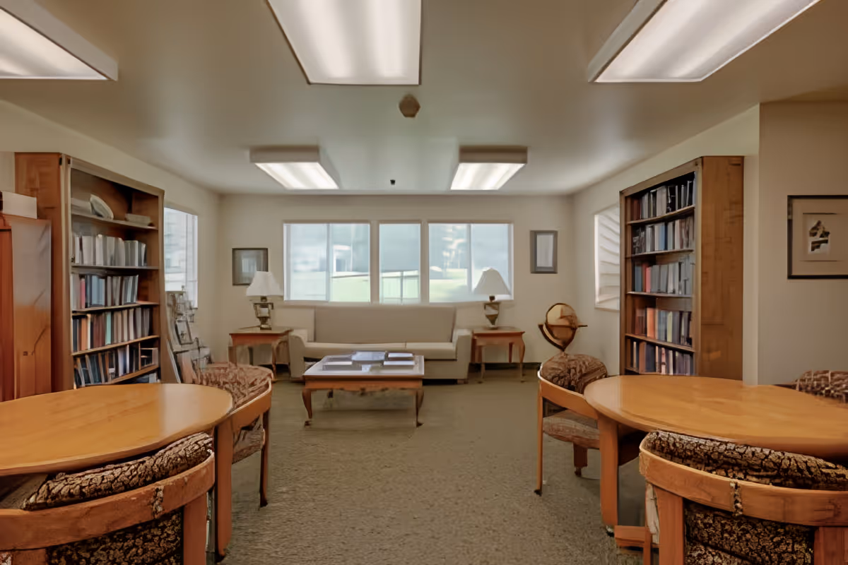 A cozy communal lounge with bookshelves, round tables and chairs, a sofa and coffee table under fluorescent lights.