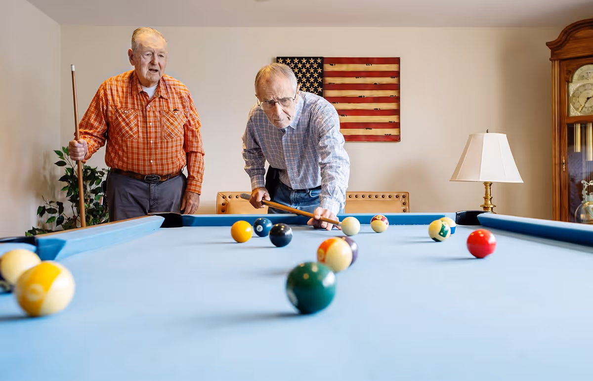Two elderly men playing pool in a room with a blue pool table. One man is aiming to take a shot while the other stands nearby holding a cue stick. An American flag is displayed on the wall behind them, and there is a lamp and a grandfather clock in the background.