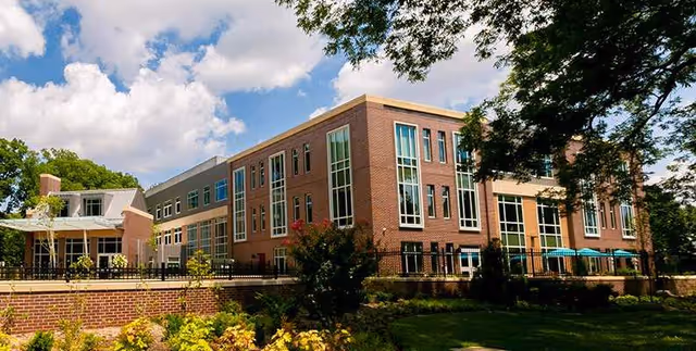 Exterior view of a multi-story brick building with large windows, surrounded by trees and greenery under a partly cloudy sky.