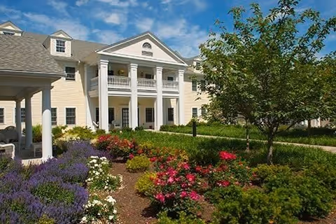 Exterior view of a large, elegant building with white columns and a balcony, surrounded by a well-maintained garden with colorful flowers and green shrubs under a blue sky with some clouds.