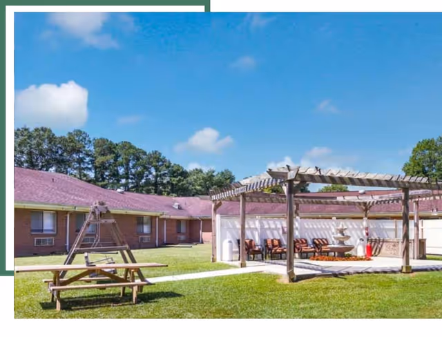 Brick building with red roof surrounds a grassy courtyard featuring a pergola and picnic area.