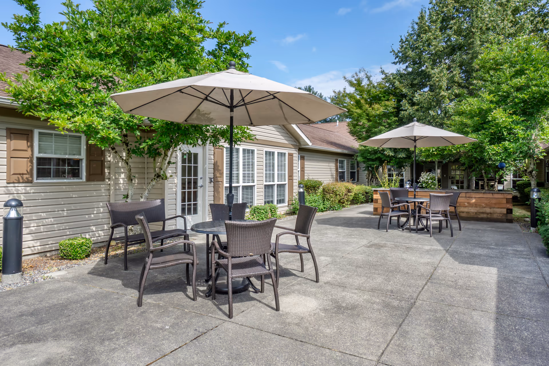 Outdoor patio area with two round tables, each surrounded by four wicker chairs and shaded by large beige umbrellas. The patio is adjacent to a beige building with windows and a door, surrounded by green trees and shrubs under a partly cloudy blue sky.