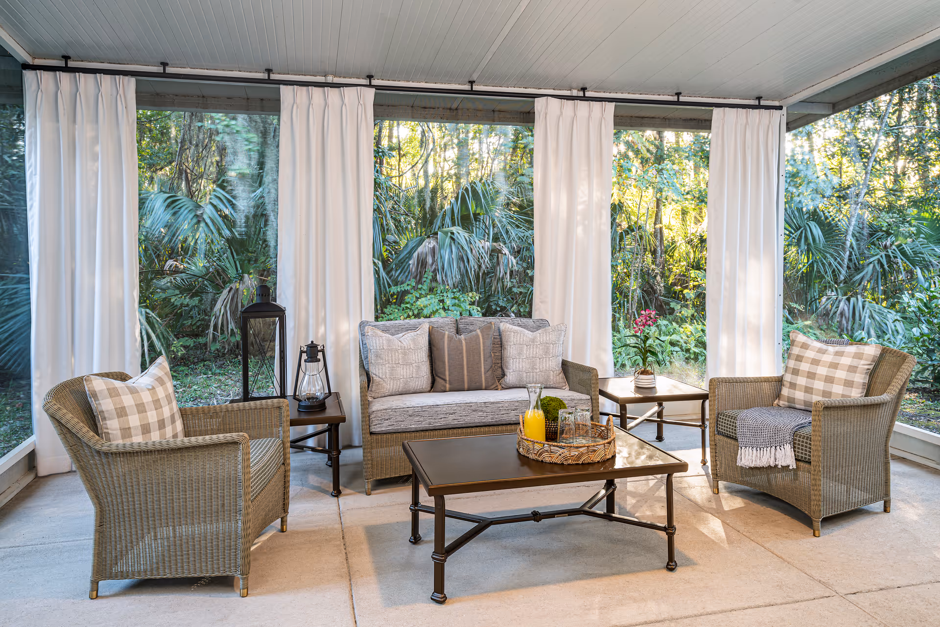 A cozy screened-in porch with wicker furniture including a loveseat and two armchairs, all with cushions and plaid pillows. There are two side tables, one with a lantern and the other with a small potted plant. A coffee table in the center holds a tray with a pitcher of orange juice and glasses. White curtains hang around the porch, and lush green foliage is visible outside.