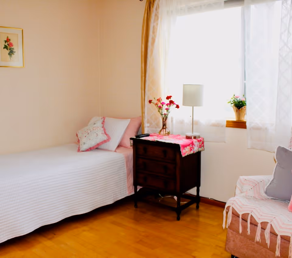 Sunlit bedroom with a single bed, a wooden nightstand holding a lamp and flowers beside a curtained window and an upholstered chair.