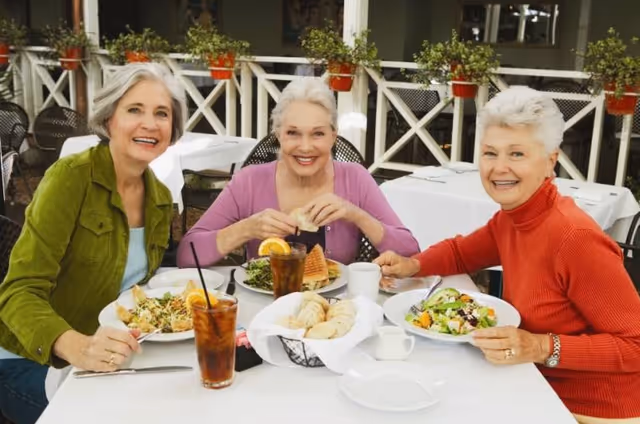 Three elderly women sitting at a table outdoors, smiling and enjoying a meal together with plates of salad, sandwiches, and iced tea on the table.
