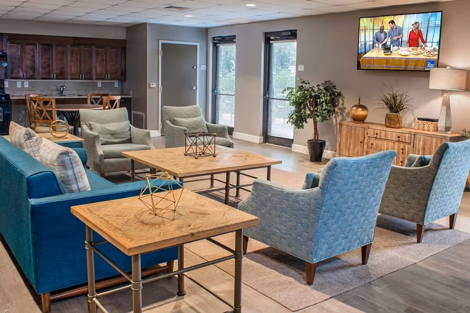 A cozy senior living facility common area with a blue sofa, four armchairs, two wooden coffee tables with decorative geometric sculptures, a wooden sideboard with plants and a lamp, and a wall-mounted TV showing a cooking show. The room has large windows and a door letting in natural light, with a kitchen area visible in the background.