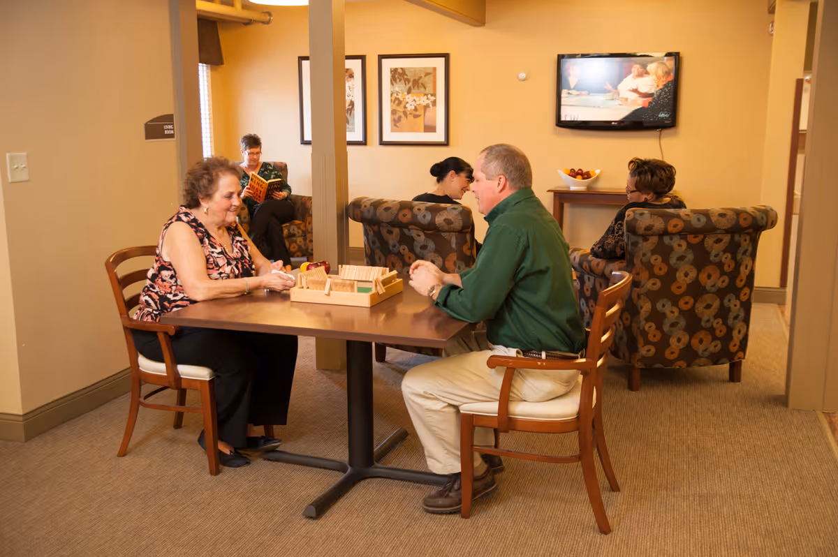 Two elderly people sitting at a table playing a board game in a cozy room. In the background, three other elderly people are seated on patterned armchairs, one reading a book and two engaged in conversation. The room has warm lighting, framed artwork on the walls, and a television mounted on the wall.