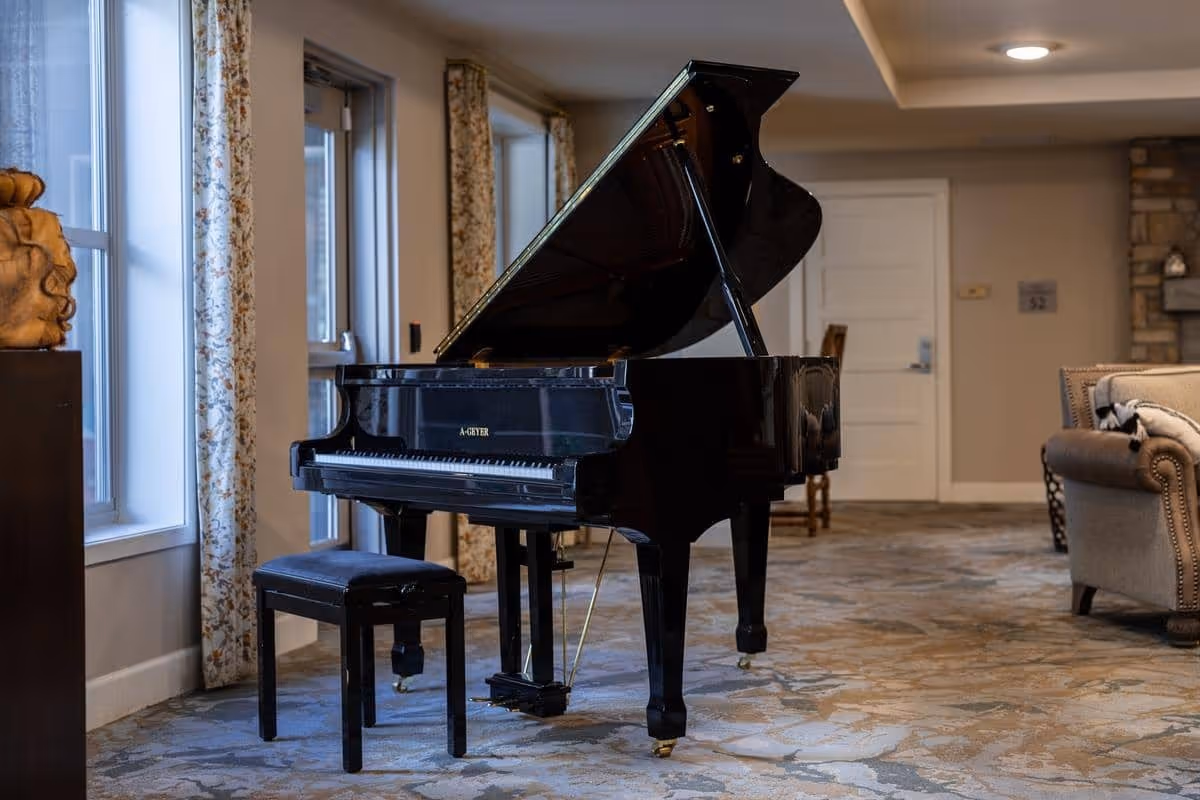 A black grand piano with an open lid and matching bench sits on a patterned carpet in a cozy room with beige walls, floral curtains, a stone fireplace, and a beige upholstered armchair.