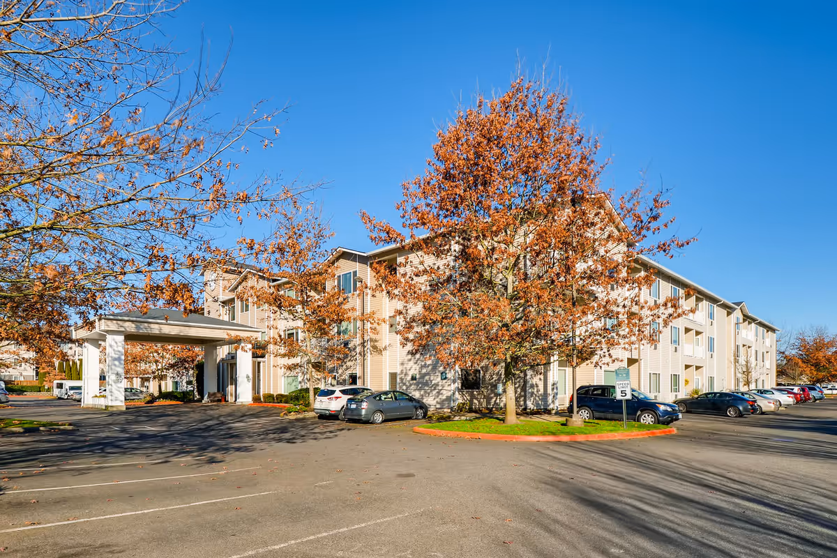 Three-story beige senior living building with a covered entrance, parked cars, and trees with autumn leaves under a clear blue sky.