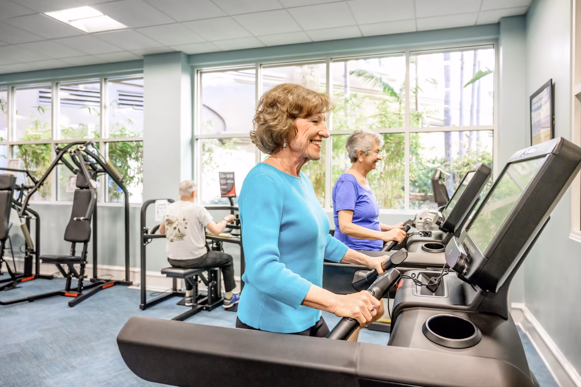 Three elderly individuals exercising in a fitness room with large windows. Two women are walking on treadmills, smiling, while a man is using a seated exercise machine in the background. The room has light blue walls and various exercise equipment.
