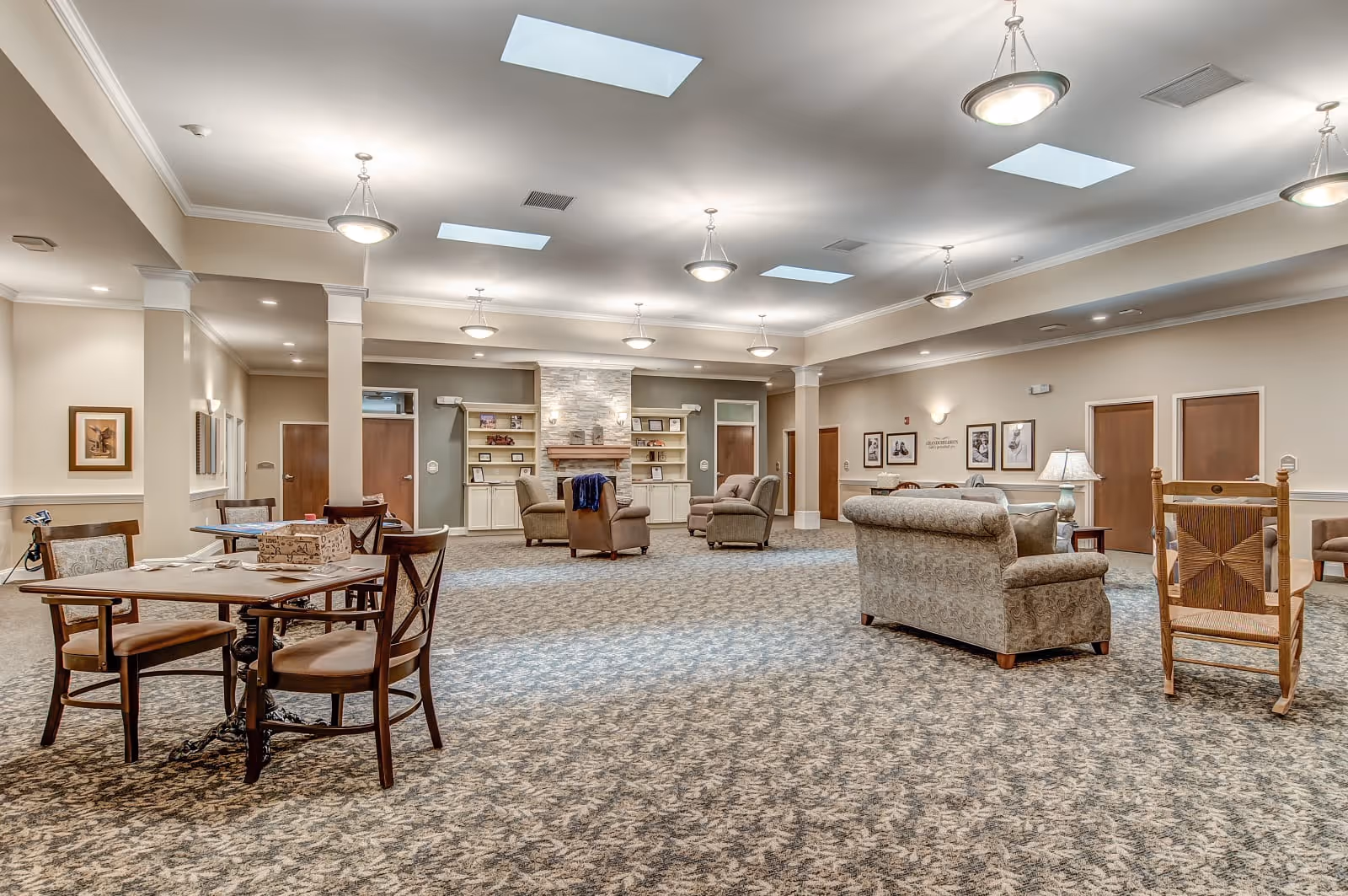 Spacious furnished common room with sofas, armchairs, tables, and a fireplace in a senior living facility.