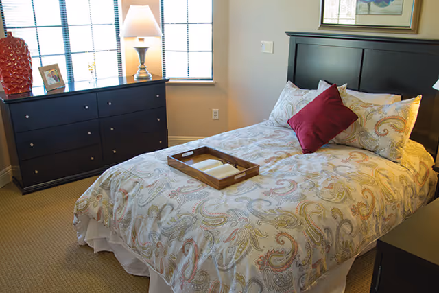 A neatly made bed with patterned bedding and a red accent pillow in a sunlit bedroom with a dresser and lamp.