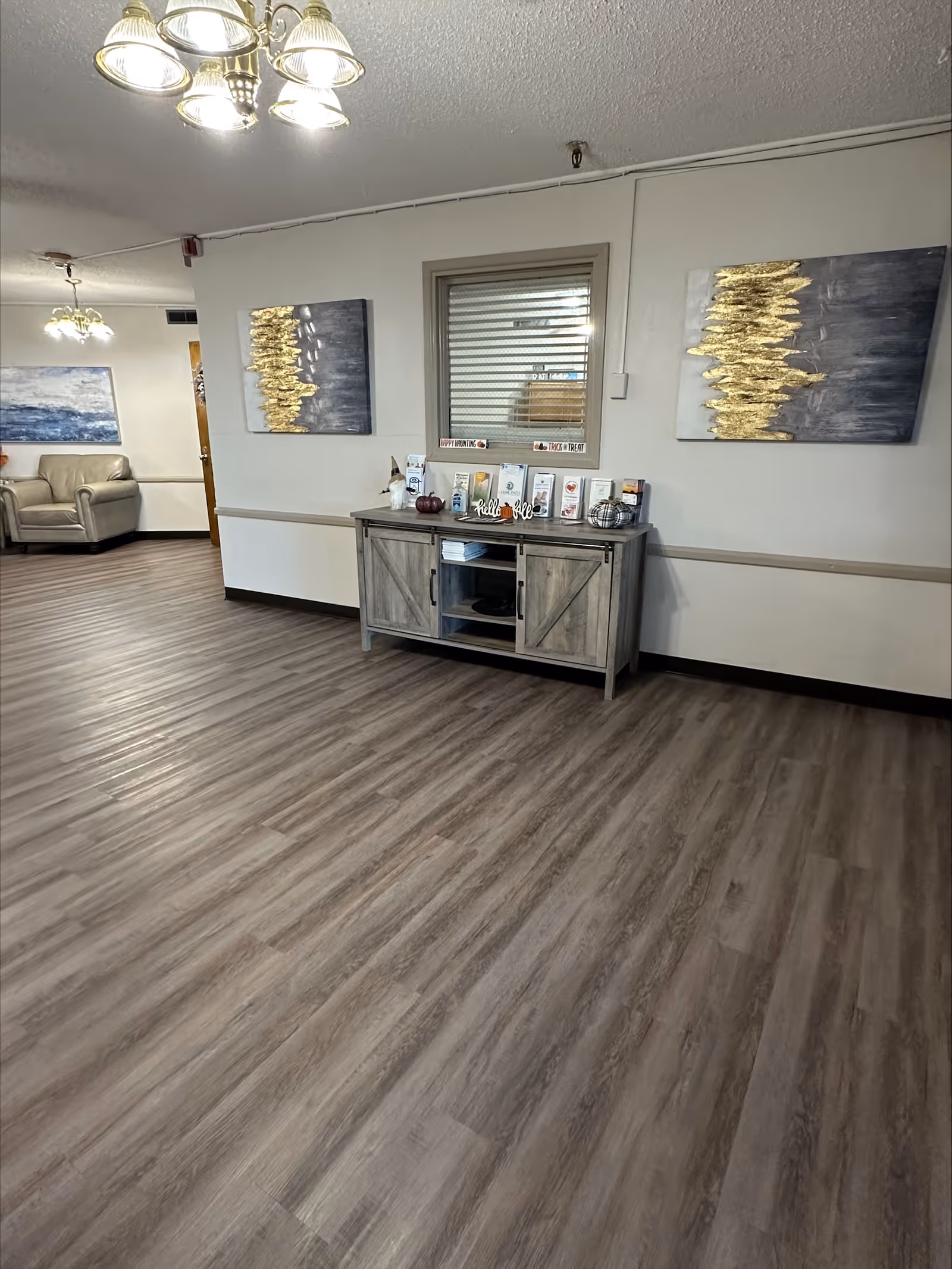 Interior view of a nursing and rehab facility hallway with wood-look flooring, a wooden cabinet against the wall displaying brochures and decorative items, two abstract paintings with gold accents on the wall, and a seating area with beige armchairs in the background.