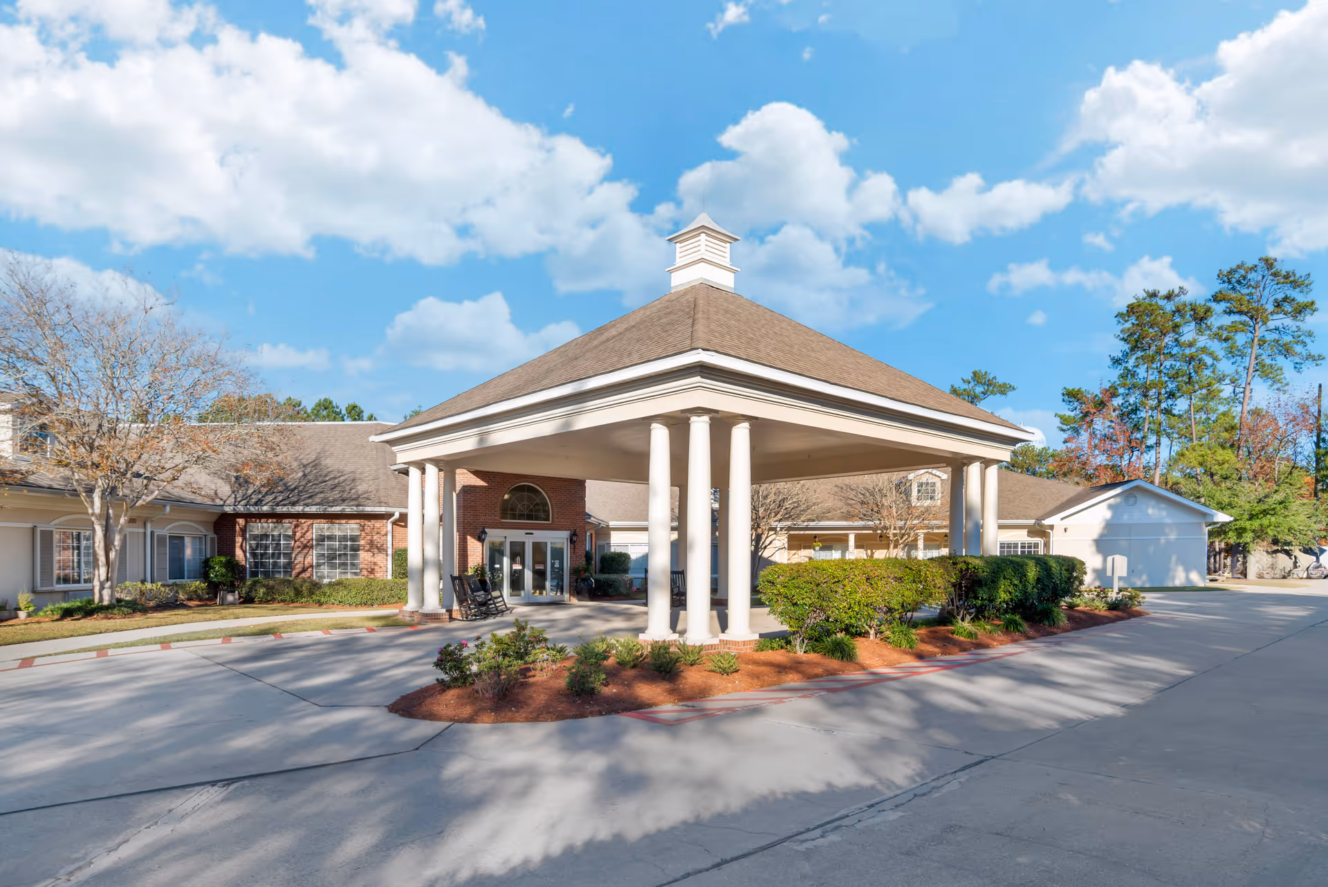 Exterior view of Brookdale Mandeville facility entrance with a covered driveway supported by white columns, surrounded by landscaped bushes and trees under a partly cloudy blue sky.
