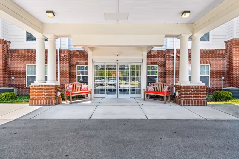 Entrance of a building with a covered porch supported by white columns and brick bases. Two wooden benches with red cushions and patterned pillows are placed on either side of the automatic glass doors. The building exterior features red brick and white siding with several windows.