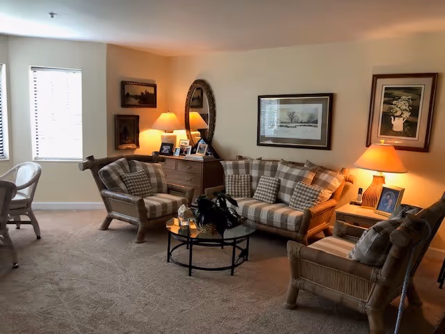 A cozy living room with beige walls and carpet, featuring a plaid sofa and two matching armchairs with wooden frames. A glass coffee table with a plant centerpiece sits in the middle. There are two table lamps on wooden side tables, framed artwork on the walls, and a wooden dresser with a round mirror and several framed photos.