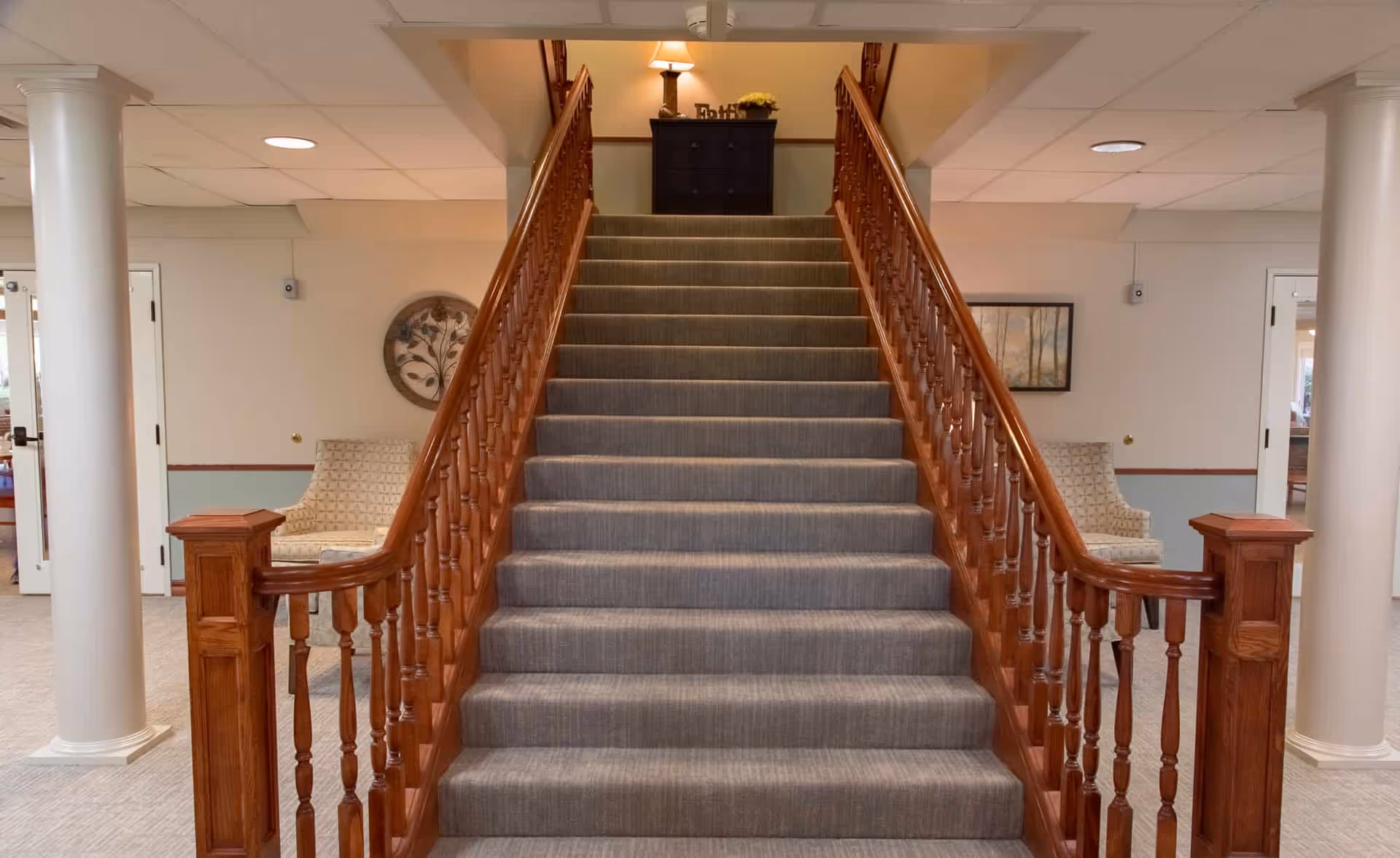 Carpeted staircase with wooden handrails in a well-lit interior space, flanked by two white columns and two patterned armchairs on either side. A small table with a lamp and decorative items is visible at the top of the stairs.