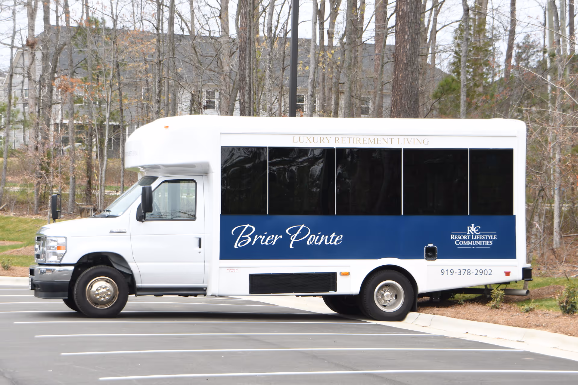 A white and blue shuttle bus parked in a parking lot with trees and residential buildings in the background. The bus has the text 'Brier Pointe' and 'Luxury Retirement Living' on its side, along with the logo and contact number for Resort Lifestyle Communities.