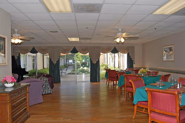 Spacious dining room with tables set with green tablecloths and red chairs, hardwood floor, ceiling fans, and large sliding glass doors opening to a courtyard.