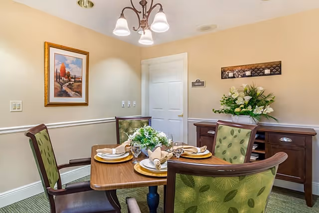 Small dining room with a wooden table set for four, green upholstered chairs, a floral centerpiece, sideboard and wall art.