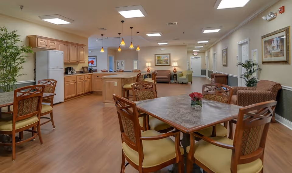 A spacious senior living common area featuring a kitchen with wooden cabinets, a white refrigerator, and a countertop with bar stools. In the foreground, there are dining tables with cushioned chairs. The room has wood flooring, warm lighting from ceiling fixtures and pendant lights, and comfortable seating areas with armchairs along the walls. Decorative plants and framed artwork adorn the space.
