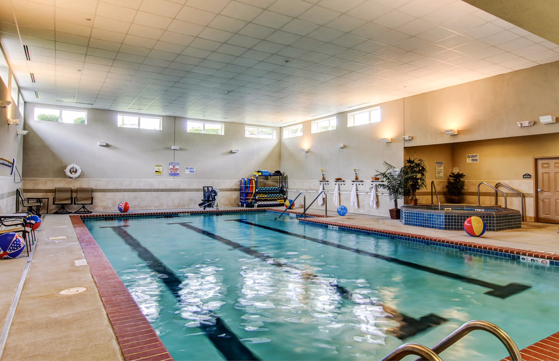Indoor swimming pool area with clear water, lane markings, and several colorful beach balls floating or placed around the pool. The room has high windows letting in natural light, beige walls, and a tiled ceiling. There is a hot tub in the corner with handrails, towels hanging on the wall, some chairs along the side, and pool safety equipment visible.