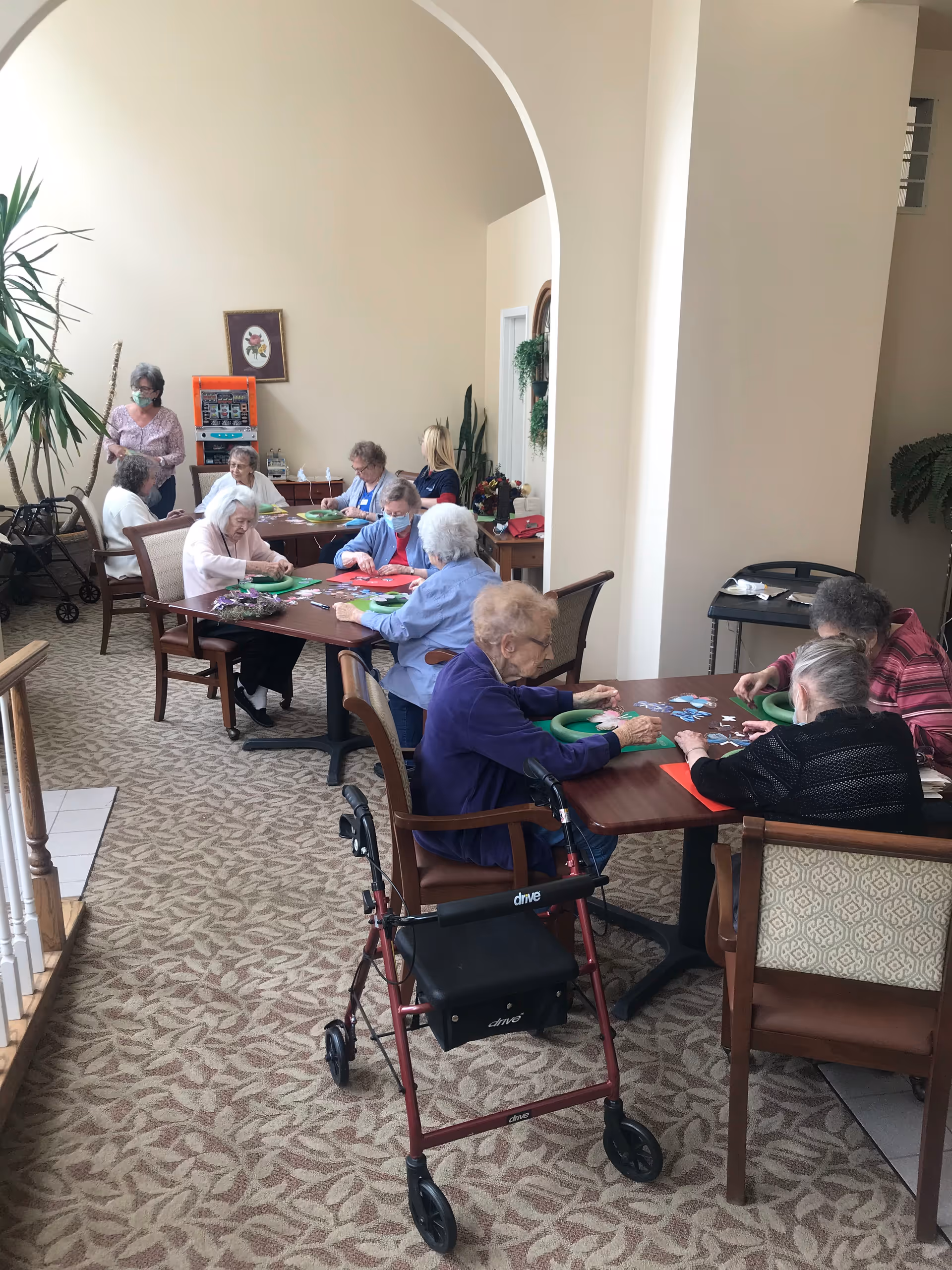 A group of elderly women sitting around tables in a well-lit room, engaging in a craft or puzzle activity. Some women are wearing masks, and there is a walker visible in the foreground. The room has beige walls, carpeted floors with a leaf pattern, and a large archway. There are plants and framed artwork on the walls.