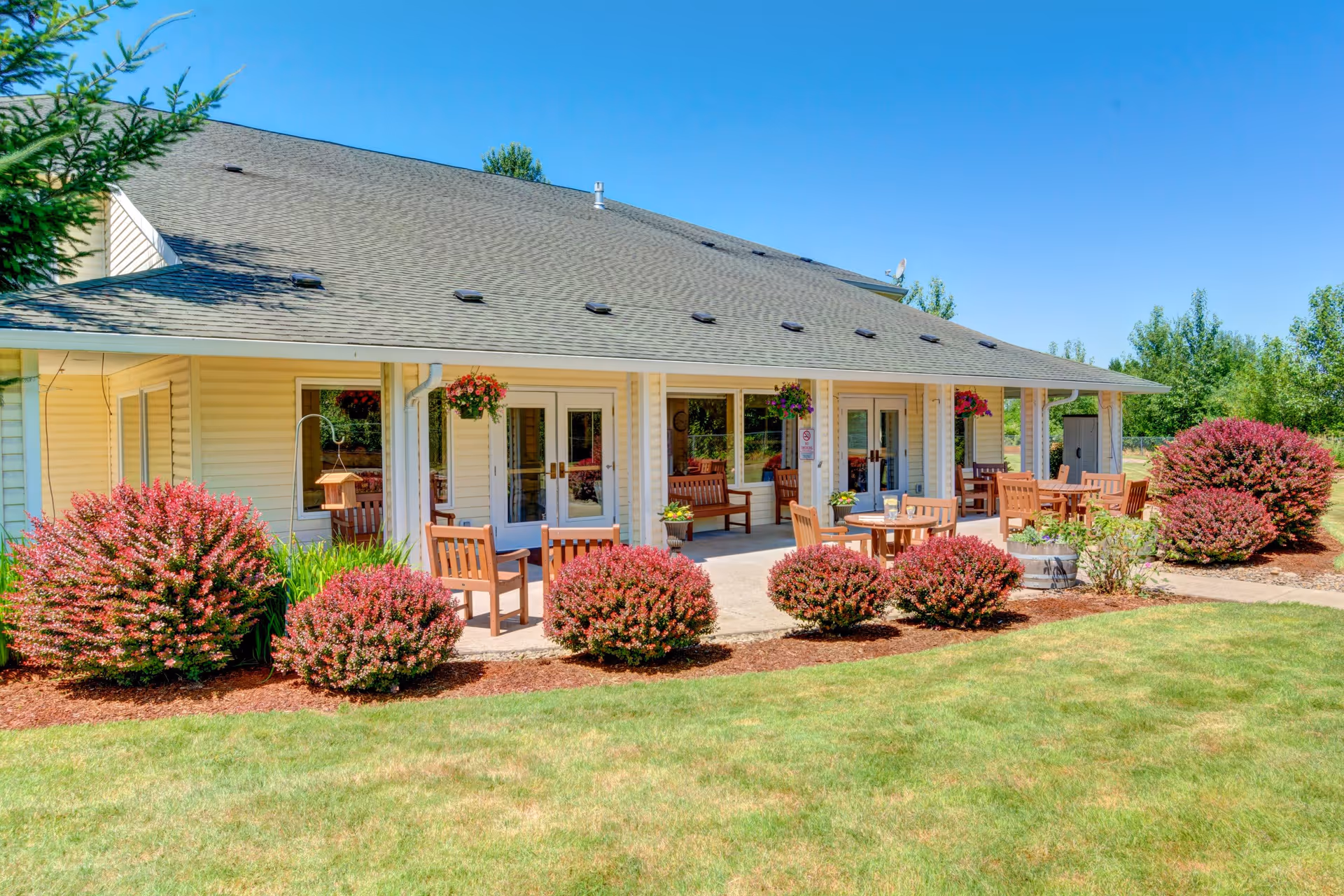 Outdoor patio area of a senior living facility with wooden chairs and tables arranged on a concrete patio. The patio is adjacent to a beige building with large windows and glass doors. There are hanging flower baskets and neatly trimmed bushes with red foliage surrounding the patio. The sky is clear and blue, and the lawn in front is green and well-maintained.