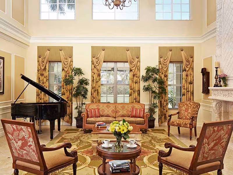 A spacious and elegant living room with high ceilings and large windows adorned with gold curtains. The room features a grand piano on the left, a patterned sofa with red and gold cushions in the center, two matching armchairs, and a round wooden coffee table with a vase of yellow flowers and two cups. There are also potted plants near the windows and a decorative fireplace on the right.