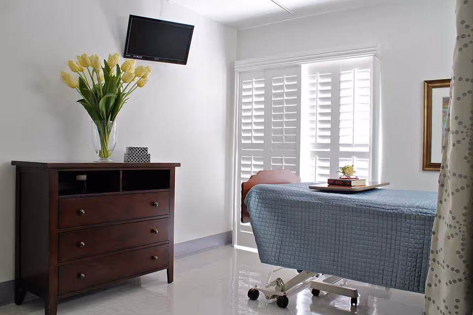 A bright room with a hospital-style bed covered in a blue quilted blanket, a wooden dresser with a vase of yellow tulips, a small TV mounted on the wall, and white plantation shutters covering the window.