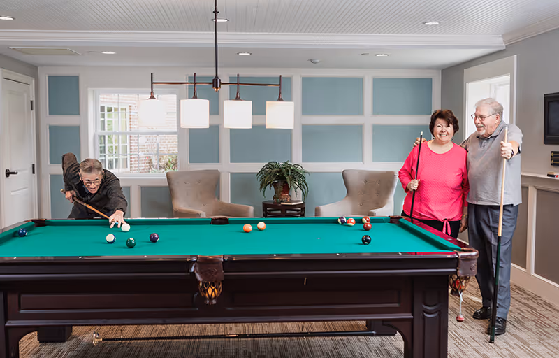Three elderly people in a well-lit room with a pool table. One person is taking a shot while the other two stand nearby smiling, holding pool cues. The room has light blue and white paneled walls, two armchairs, a small table with a plant, and a window letting in natural light.