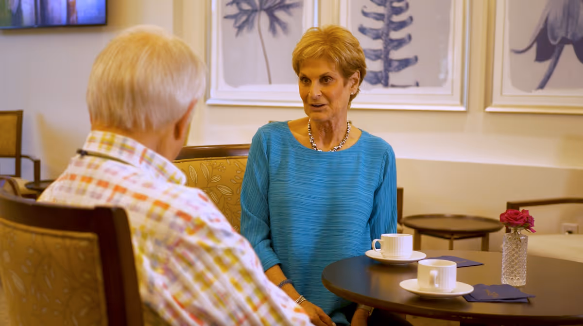 Two elderly individuals sitting at a round table in a cozy indoor setting, engaged in conversation. The table has two white coffee cups on saucers, a small vase with a pink flower, and two dark blue napkins. The background features framed botanical artwork on the wall and comfortable chairs.