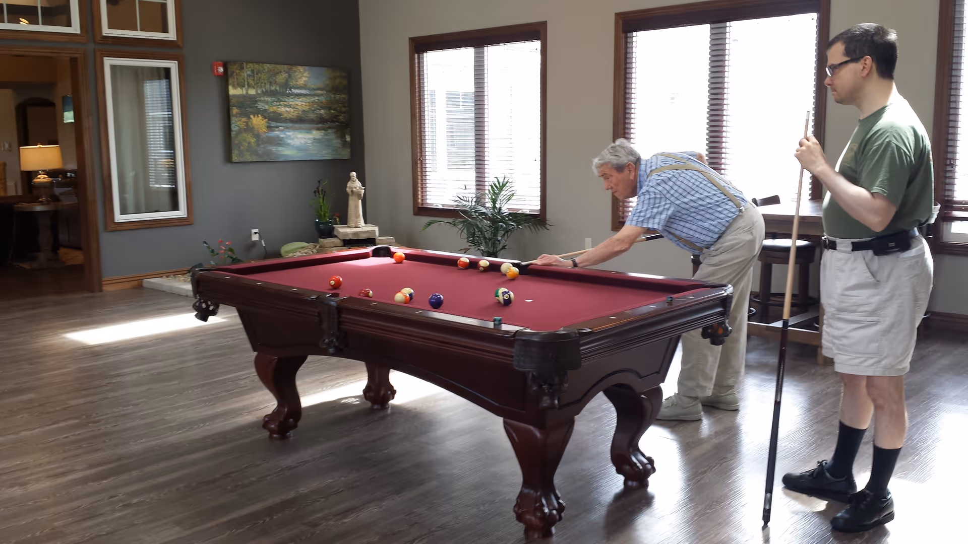 Two men playing pool at a burgundy-felt billiards table in a bright common room with wood floors and large windows.